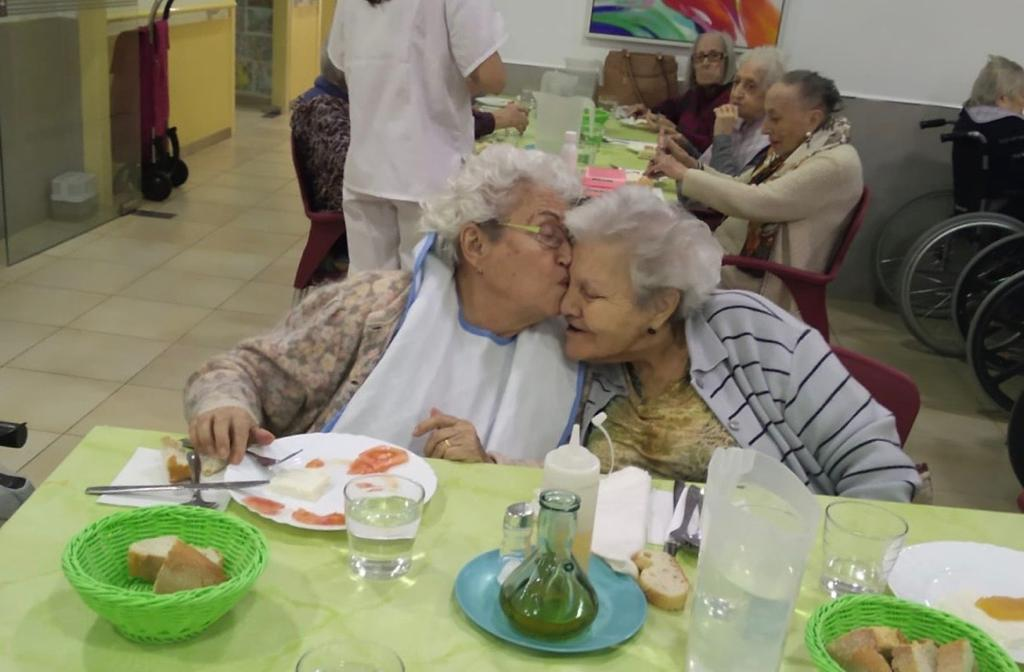 Dos señoras mayores sentadas a un mesa. Una le está dando un beso en la mejilla a la otra.